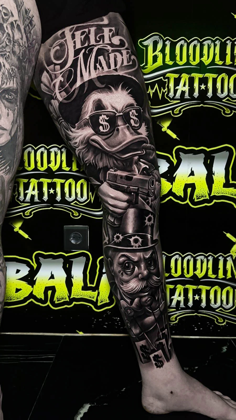 Detailed black and grey Chicano leg sleeve tattoo featuring Scrooge McDuck, Monopoly Man, cash-sign glasses, and bullet hole accents at Bloodline Tattoo Bali.