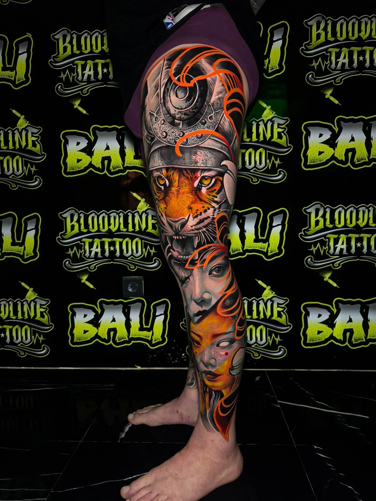 Colour leg sleeve tattoo with a roaring tiger in a samurai helmet, soft female portraits, and orange ornamental swirls at Bloodline Tattoo Bali.