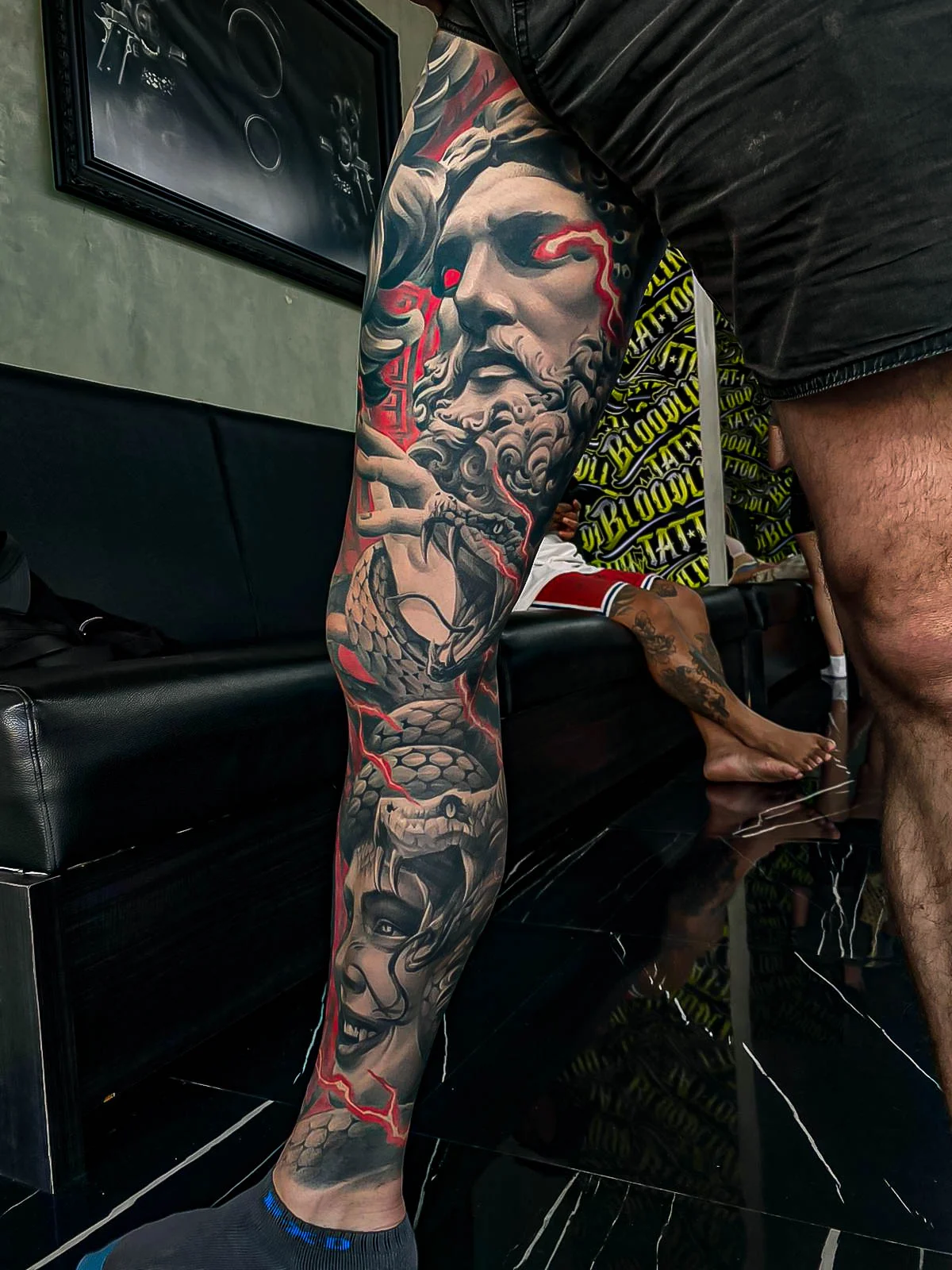 Healed black-and-grey leg sleeve with a bearded god, Medusa face, and snake scales, lifted by red lightning accents and crisp healed detail at Bloodline Tattoo Bali.
