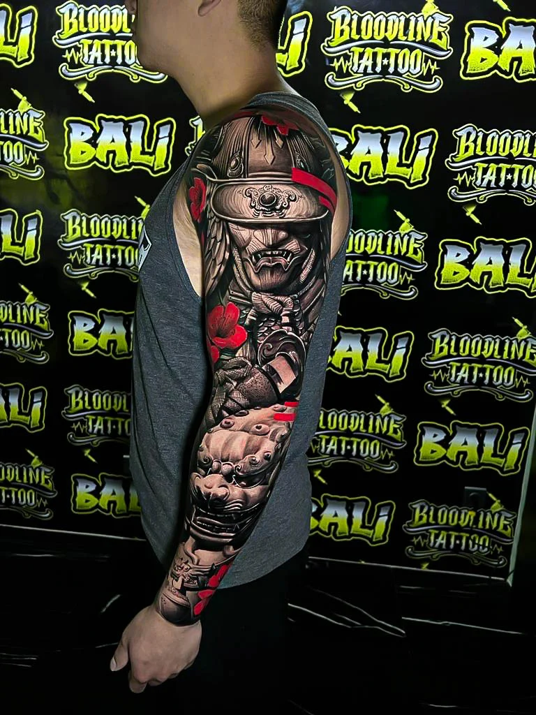 Black and grey Japanese full sleeve with a samurai kabuto, armor details, cloud shading, and red flower accents at Bloodline Tattoo Bali.