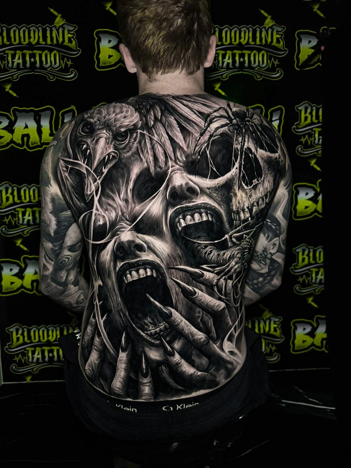 A black and grey realism full back piece with a cracked skull, screaming faces, and dark horror shading spread across the shoulder blades, at Bloodline Tattoo Bali.