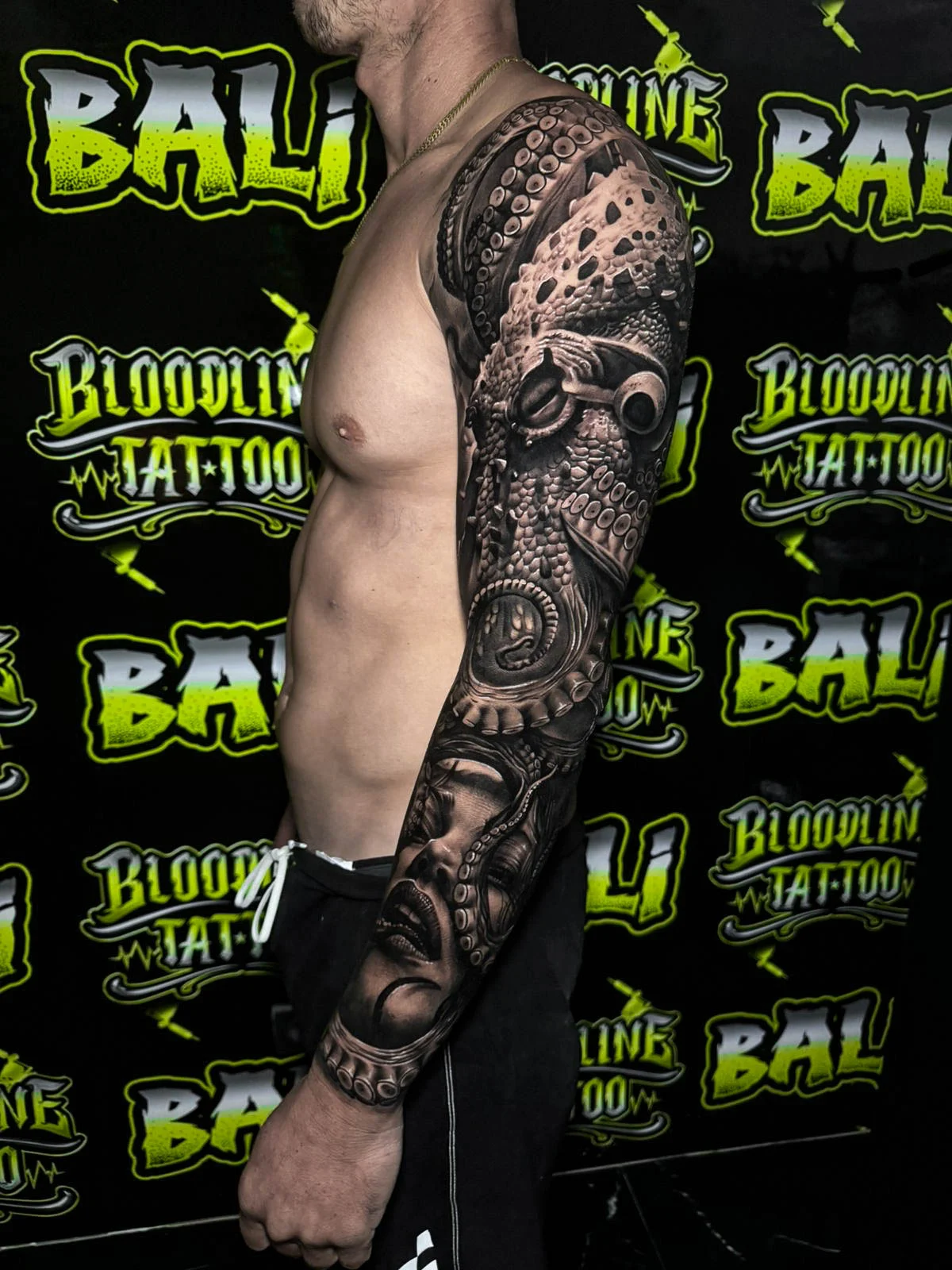 A black and grey realism half sleeve with clockwork detail, ornamental patterns, and dark portrait elements wrapping the upper arm, at Bloodline Tattoo Bali.