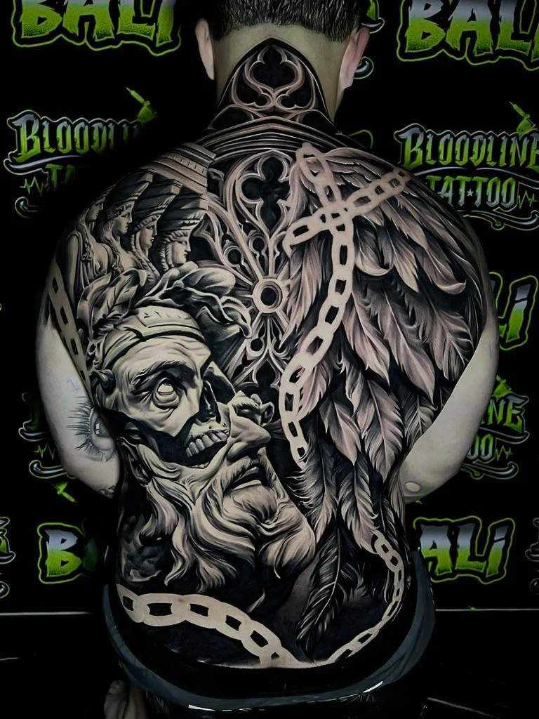 A black and grey realism full back piece with an aged warrior face, a skull headdress, and feathered textures in bold black and grey contrast, at Bloodline Tattoo Bali.