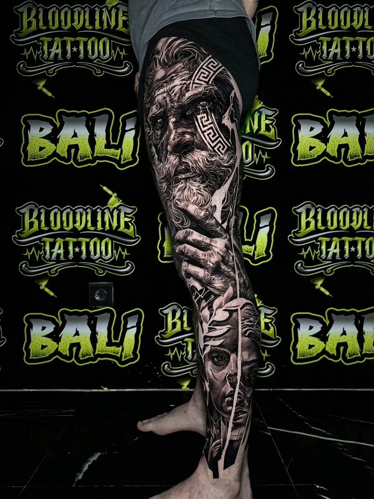 A black and grey realism lower leg sleeve with a stern elder face, feminine portrait detail, and ornamental linework in deep shadows, at Bloodline Tattoo Bali.